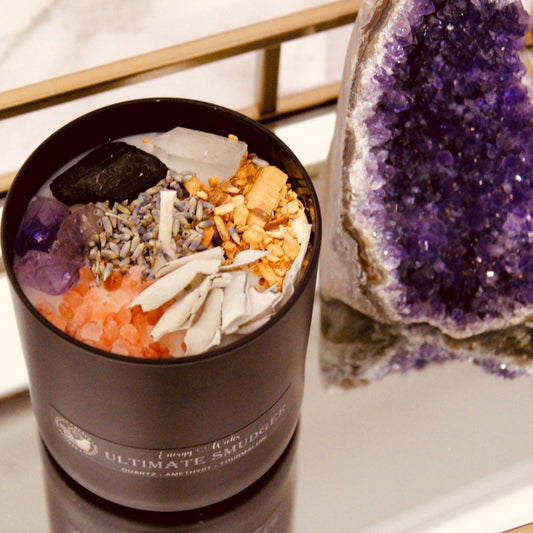 6oz soy candle with one wick, nestled in a black container next to amethyst pillar. Adorned with dried lavender, Black Tourmaline, Amethyst, and clear Quartz crystals, it carries a Basil, Mint, and Sage fragrance. Part of the Ultimate Smudger crystal candle collection, designed to cleanse spaces and clear negative energies.